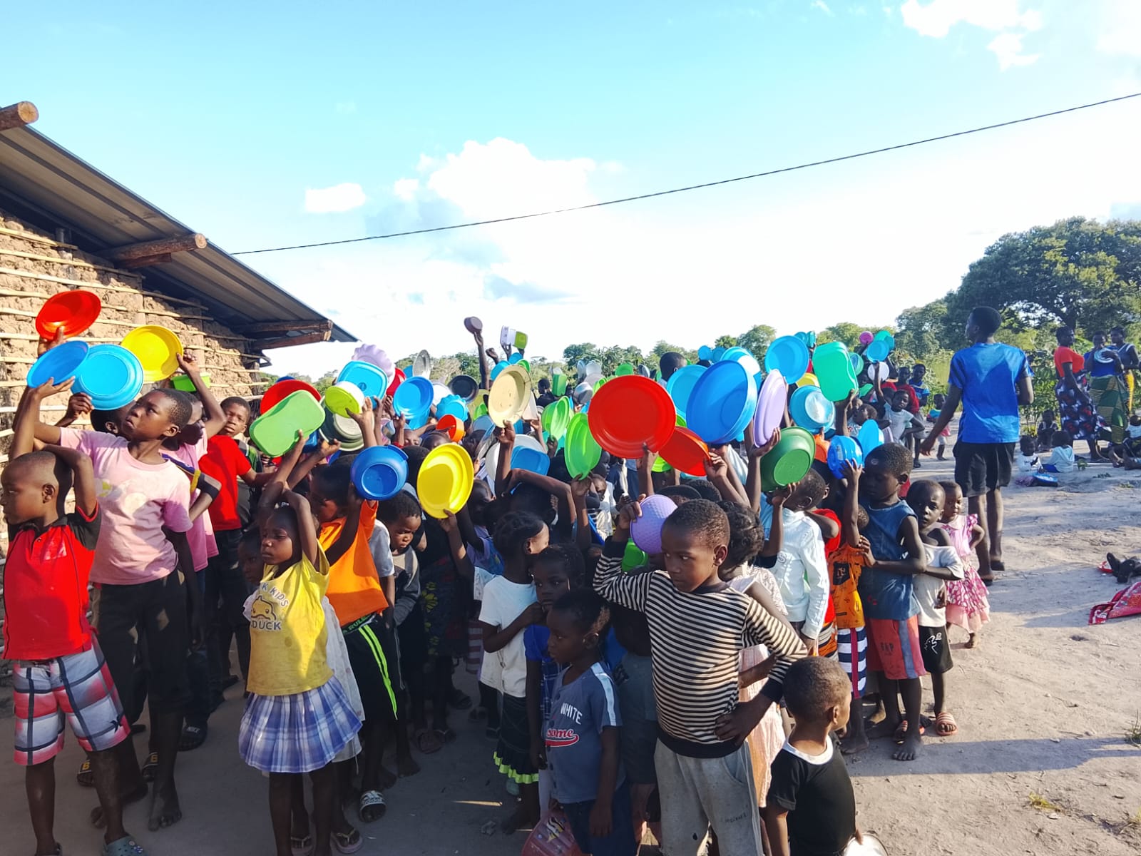 Multidão de crianças em Moçambique levantando pratos coloridos durante a distribuição de alimentos.
